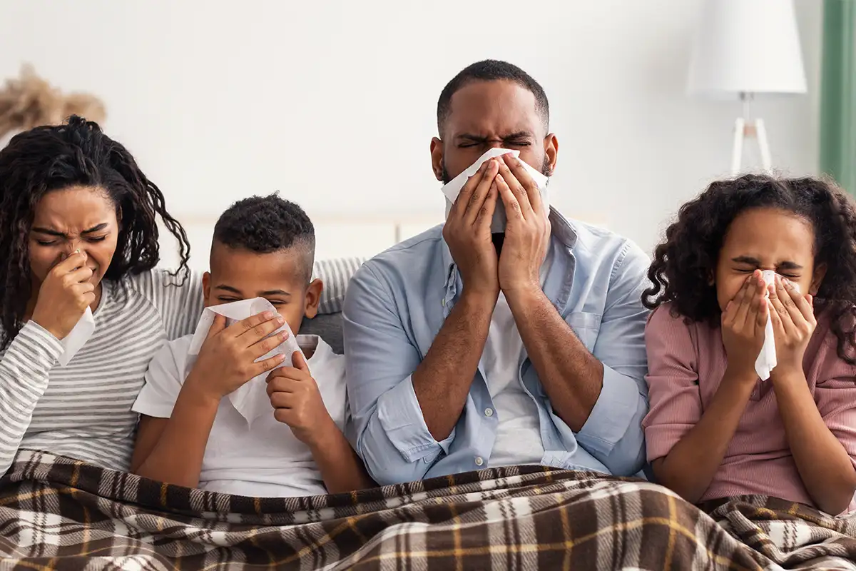 a family of four sick on the couch holding tissues to their faces