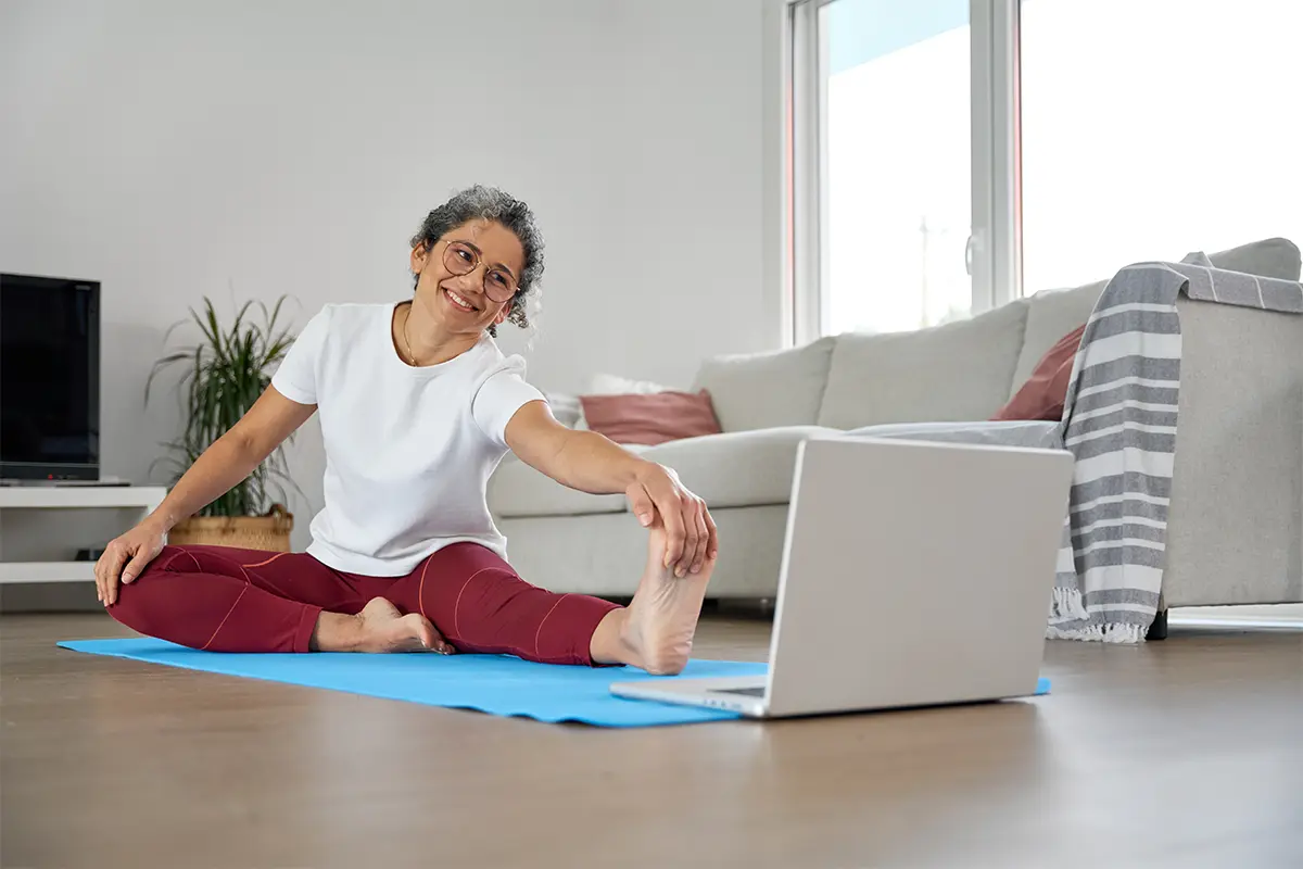 a woman stretches on the floor while watching a laptop during a virtual exercise class